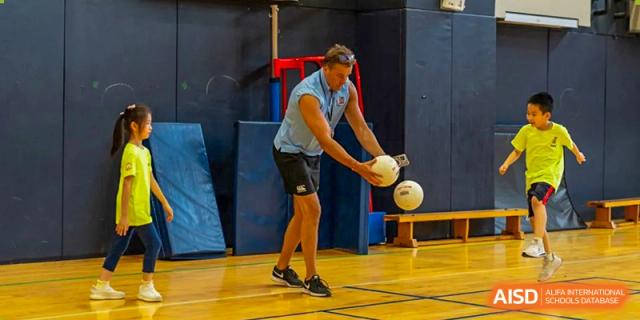 A male teacher or coach from Wellington College International Tianjin is teaching a student how to handle a ball during a sports lesson.