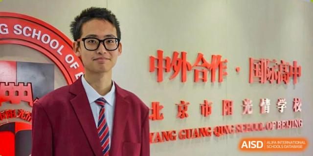 A male student in a red blazer and tie standing in front of the school logo at Yang Guang Qing International School of Beijing.