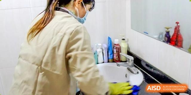 A staff member wearing gloves and a mask cleaning a sink and counter in a restroom or dormitory at Yang Guang Qing International School of Beijing.