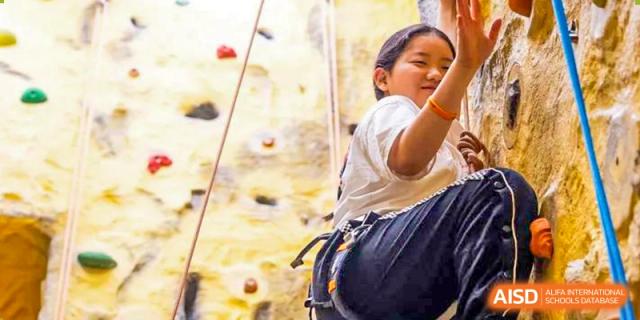 Huili School Shanghai student rock climbing indoors, secured by a harness and rope, looking up at the wall.
