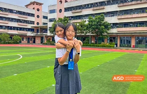 Two female students at Shenzhen Foreign Languages GBA Academy smiling and posing for a picture on a green field, one giving the other a hug from behind.