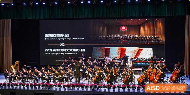 A live symphony orchestra performing on a stage at Shenzhen Foreign Languages GBA Academy, with the screen displaying the event title.