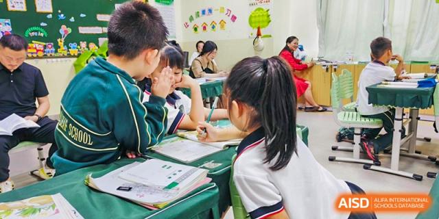 A female student from Maple Leaf International Academy Shenzhen is raising her hand during class to ask a question while other students are looking at their books.