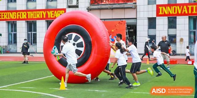 Students from Maple Leaf International Academy Shenzhen are pushing a large inflatable red ring on a sports field.