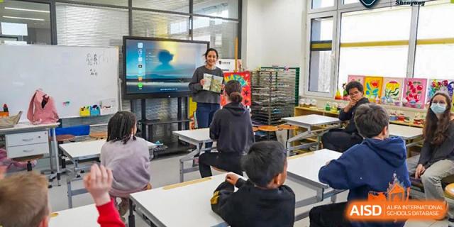 A teacher at HuZ Deutsche Schule Shenyang reading to students in a classroom with a large screen.