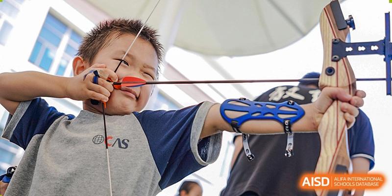 A young boy from Overseas Chinese Academy of Chiway Suzhou practicing archery, smiling as he holds a bow and arrow.