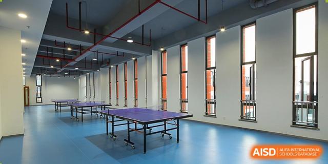 A large, empty indoor sports area at Overseas Chinese Academy of Chiway Suzhou with a blue floor and a table tennis table.