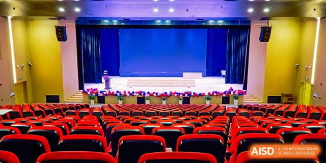 A large, empty auditorium at Overseas Chinese Academy of Chiway Suzhou with rows of red chairs facing a stage.