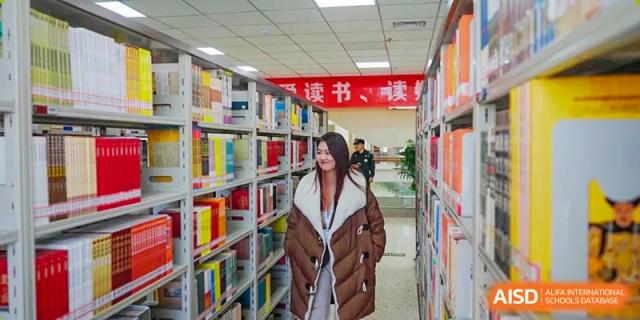 Student wearing a puffy coat walks down an aisle lined with colorful books in the library at Shanxi Plinth International School.