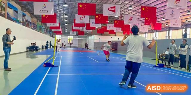 Students playing badminton or a similar sport in a large indoor gymnasium decorated with Chinese flags and banners at Shanxi Plinth International School.