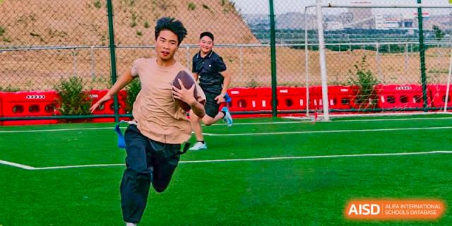 Student running while holding an American football outdoors on a turf field at Shanxi Plinth International School.