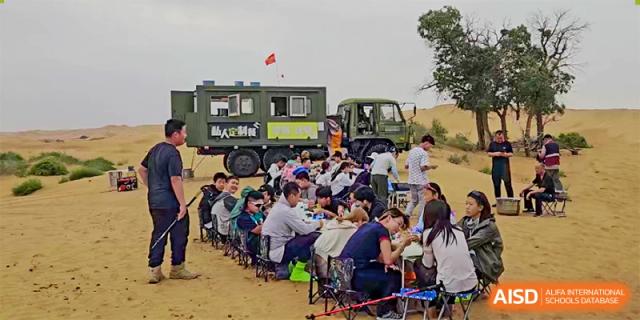 An outdoor group meal setup with participants eating at long tables beside a large utility truck, part of a Shanxi Plinth International School activity.