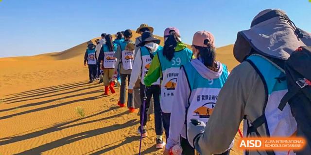 Students and participants lined up and walking with trekking poles in a sunny desert landscape, wearing matching vests for the Shanxi Plinth International School challenge.