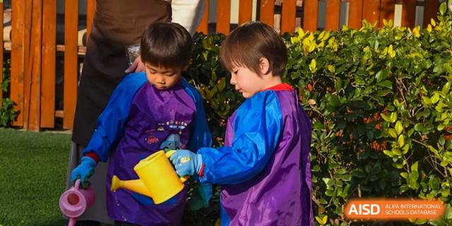 Two young children watering plants outdoors with a yellow can at Wuxi Boston International School.