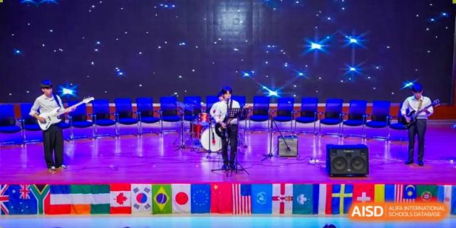 A student from Wuxi Boston International School performing an electric guitar solo on a stage, with a drum set, microphone stands, a starry background, and a line of international flags below the stage.