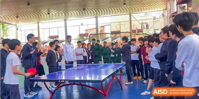 Students and teachers from Wuhan Britain-China School gather around a ping pong table during a recreational activity in an open sports area.