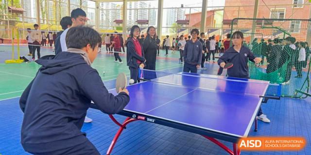 Two people playing table tennis (ping pong) with spectators from Wuhan Britain-China School watching in the background.