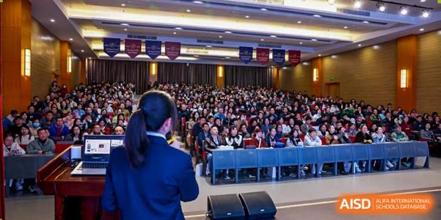 A large school assembly or presentation in a hall with a speaker addressing a large crowd of students from Wuhan Britain-China School.