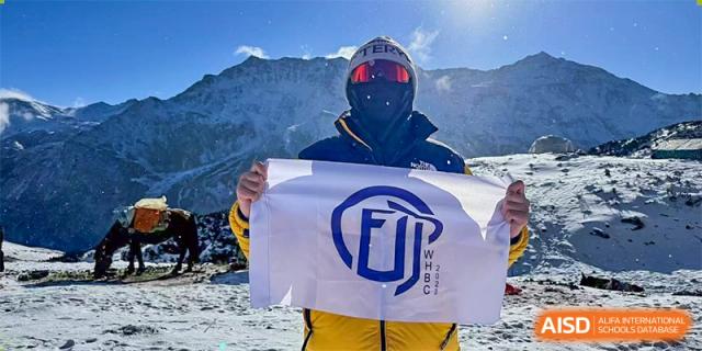 A person in winter gear holds a white flag with a blue and white logo on a snowy mountain, representing an achievement or event supported by Wuhan Britain-China School.