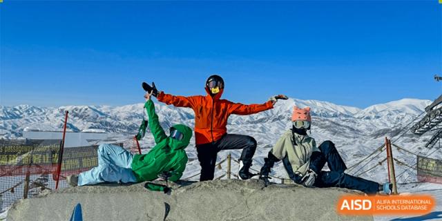 A person in an orange jacket and ski goggles poses triumphantly on a sunny, snow-covered mountain peak, a possible outdoor education trip from Wuhan Britain-China School.