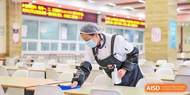 A cafeteria staff member wearing a mask and apron cleans a white table, highlighting the focus on health and hygiene at Wuhan Britain-China School.