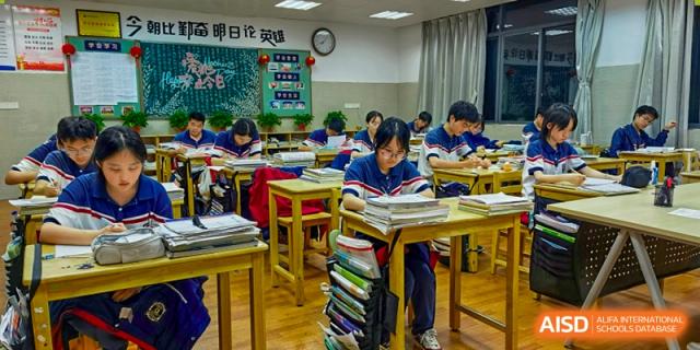 A student focused on reading or writing at a desk with a large stack of textbooks and notebooks next to them, at Ningxiang Country Garden School.