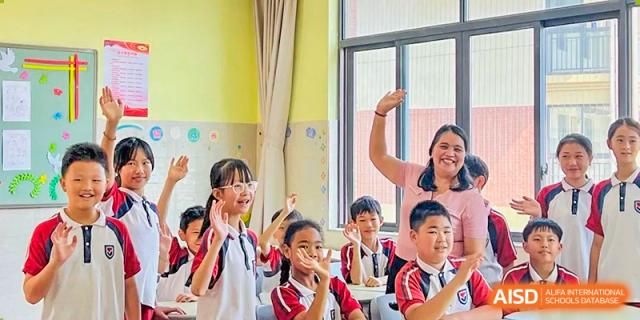 A group of cheerful primary school students smiling and raising their hands in an interactive, sunlit classroom with their teacher, at Ningxiang Country Garden School.