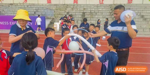 Students outdoors playing a cooperative game, holding twisted blue balloons or rings around a large white ball, with other students watching in the background, at Ningxiang Country Garden School.