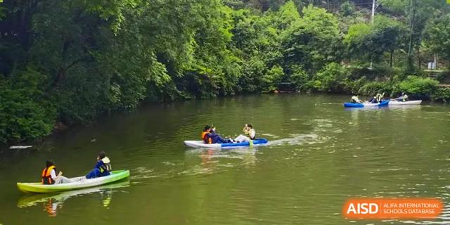 Two people in a kayak or small boat on a calm, green river or lake surrounded by trees, at Ningxiang Country Garden School.