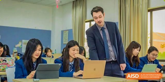 A teacher in a suit is assisting a female student working on a MacBook laptop during class time at Changsha WES (Bilingual) Academy.