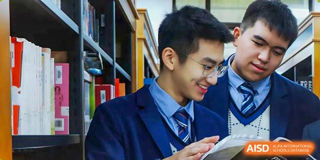 A male high school student wearing glasses and a school uniform smiling while working on a device in the school library at Changsha WES (Bilingual) Academy.