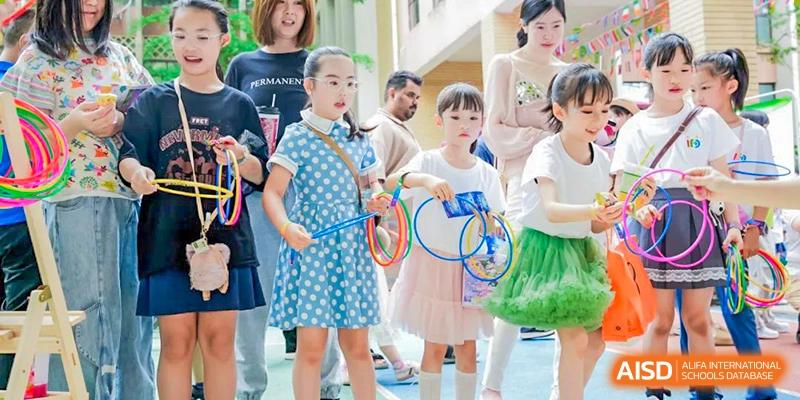 Three young girls in a crowd at a school event, the middle girl holding a racket and the other two holding colorful plastic rings at Changsha WES (Bilingual) Academy.