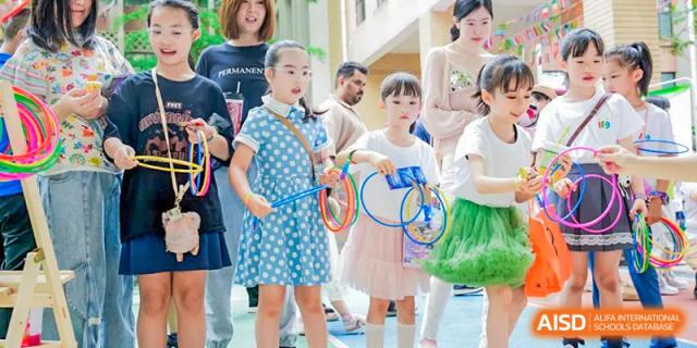 Three young girls in a crowd at a school event, the middle girl holding a racket and the other two holding colorful plastic rings at Changsha WES (Bilingual) Academy.
