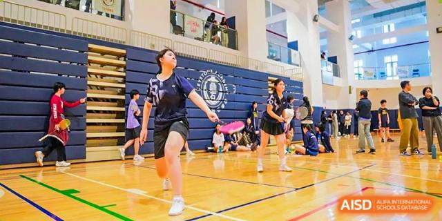 A female student jumping on a wooden gymnasium floor, holding a purple and pink racket while playing an indoor sport like badminton or pickleball at Changsha WES (Bilingual) Academy.