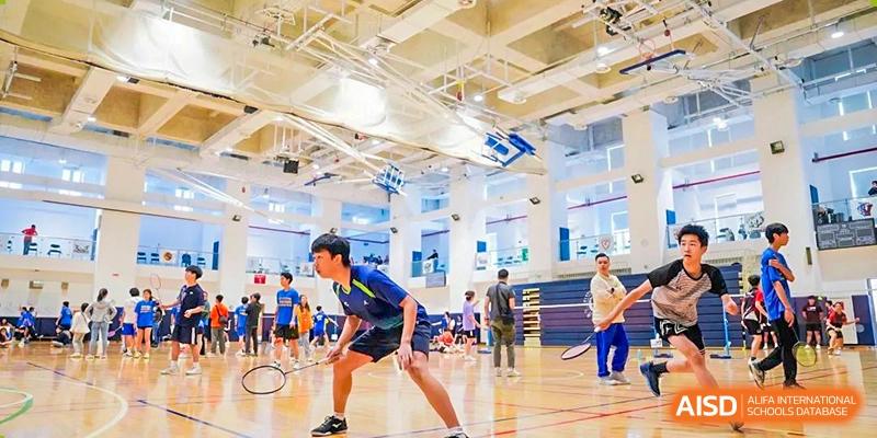 A male student in a blue and yellow uniform playing badminton aggressively on a court in a large, brightly lit gymnasium at Changsha WES (Bilingual) Academy.