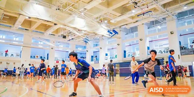 A male student in a blue and yellow uniform playing badminton aggressively on a court in a large, brightly lit gymnasium at Changsha WES (Bilingual) Academy.