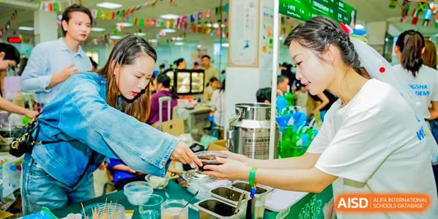 A close-up of a female student or parent serving food, likely a dessert or treat, at a busy school fair or cultural event at Changsha WES (Bilingual) Academy.