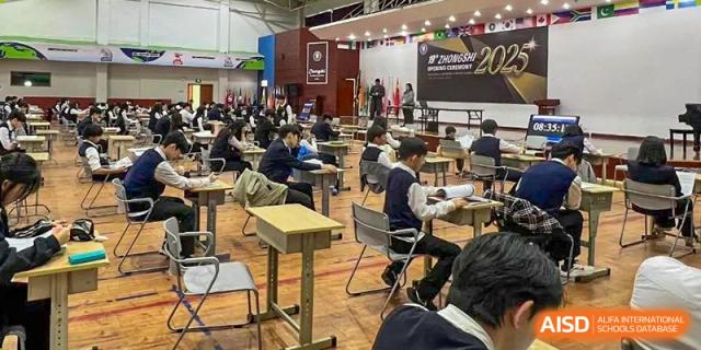 Students sitting at desks in a large multi-purpose hall or auditorium during an exam or assembly at Weihai Zhongshi Foreign School (WZFS).