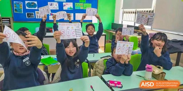 Young students in a classroom proudly holding up signs with "SOS" written on them at Weihai Zhongshi Foreign School (WZFS).