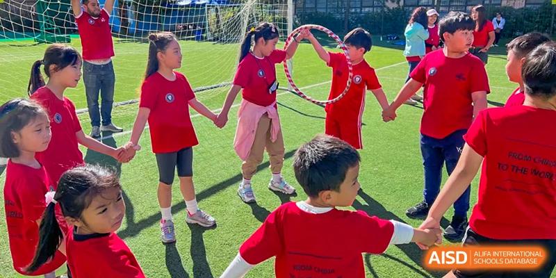 Young students at Weihai Zhongshi Foreign School (WZFS) playing an outdoor game involving a hula hoop and holding hands on a green turf.