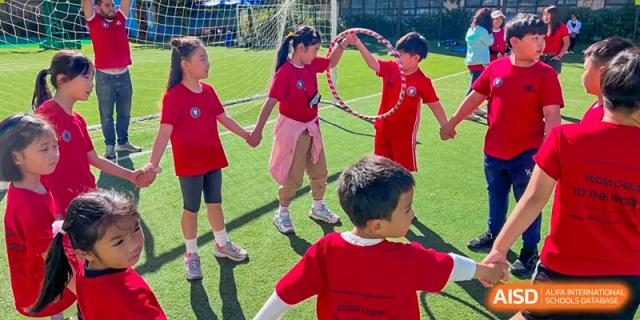 Young students at Weihai Zhongshi Foreign School (WZFS) playing an outdoor game involving a hula hoop and holding hands on a green turf.