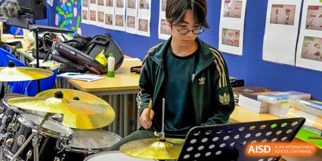 A male student wearing glasses, playing a drum set in a music room at Alcanta International College.