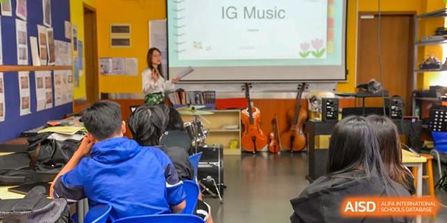 A music classroom with students and various instruments, including a cello and guitar, with a sign for "IG Music" in the background at Alcanta International College.