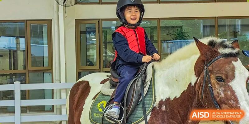 A young, smiling student from Changsha BeeMee Forest School (CBFS) wearing a helmet and red vest while riding a pony or horse.