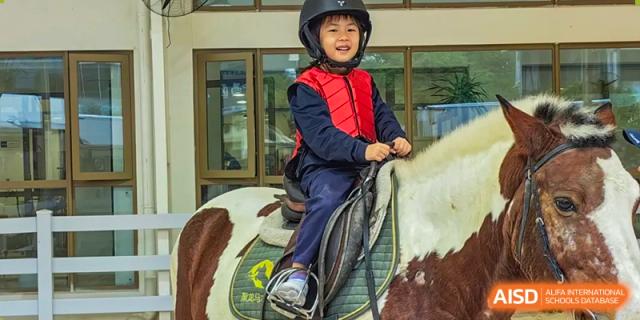 A young, smiling student from Changsha BeeMee Forest School (CBFS) wearing a helmet and red vest while riding a pony or horse.