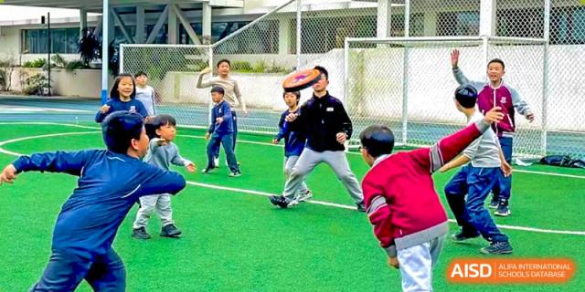 A teacher or coach from Changsha BeeMee Forest School (CBFS) playing football with young students on a small outdoor turf field.