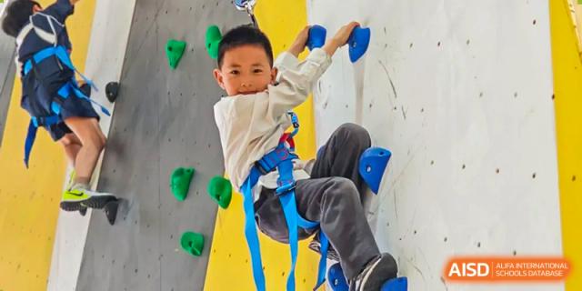 A young student from Changsha BeeMee Forest School (CBFS) wearing a safety harness and climbing a colorful indoor rock wall.