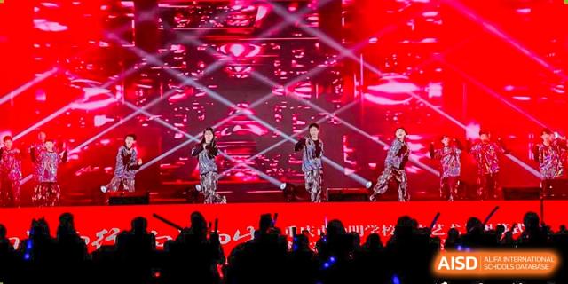 A student or performer from CHONGQING WEIMING SCHOOL singing on a stage with bright red and black lighting during a school event or concert.