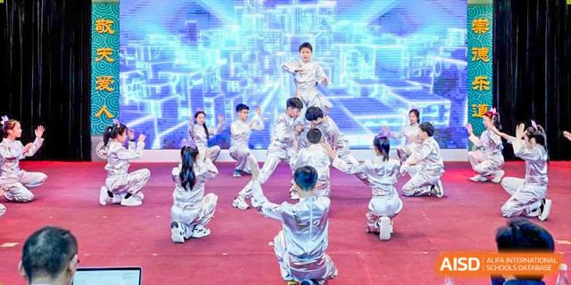 Group of students from CHONGQING WEIMING SCHOOL performing a synchronized dance routine on a stage with vibrant blue, futuristic lighting and background graphics.