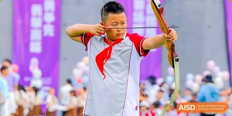 CHONGQING WEIMING SCHOOL student competitor focusing on an archery target, holding a bow and arrow during a sports competition.